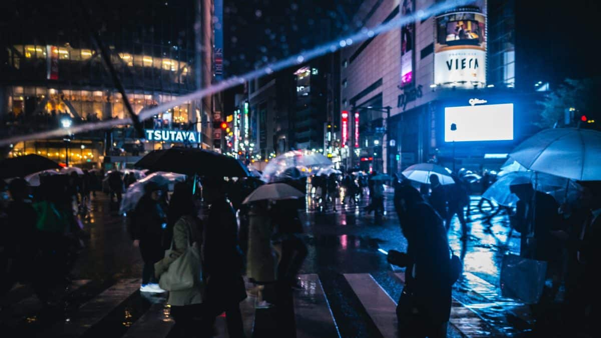 Pedestrians with umbrellas crossing a rain-soaked Tokyo street at night, neon signs reflected on the pavement