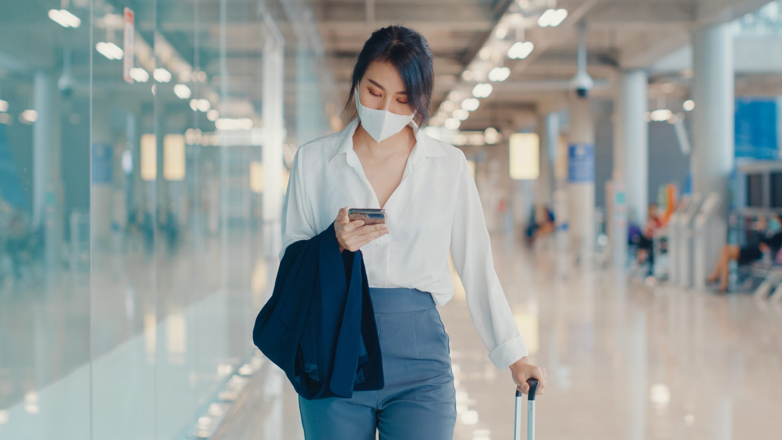 Traveler checking her phone while walking through an airport terminal with a suitcase
