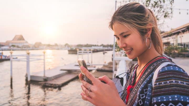 Traveler using her mobile phone by the Chao Phraya River in Bangkok at sunset