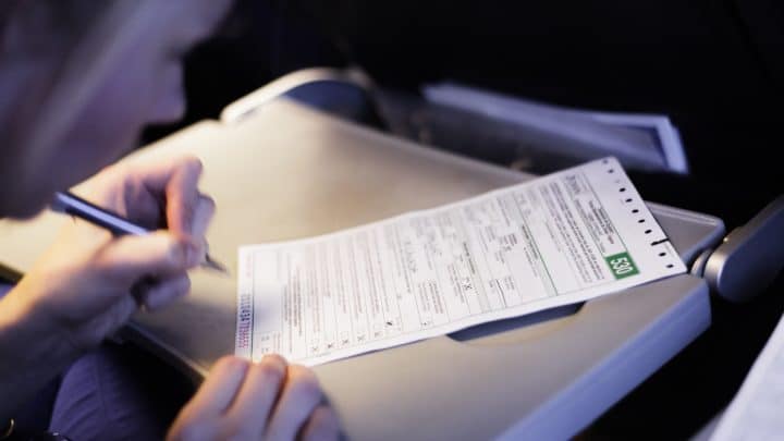 A traveler's hand filling out Japan's paper disembarkation and customs declaration card with a pen at an airplane seat tray table