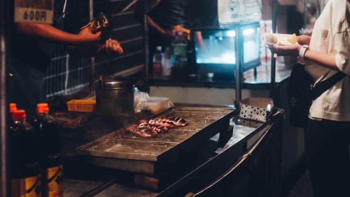 A lit-up food stall at a Japanese summer festival at night, the kind of spot where visitors stand to eat rather than walking away with the food