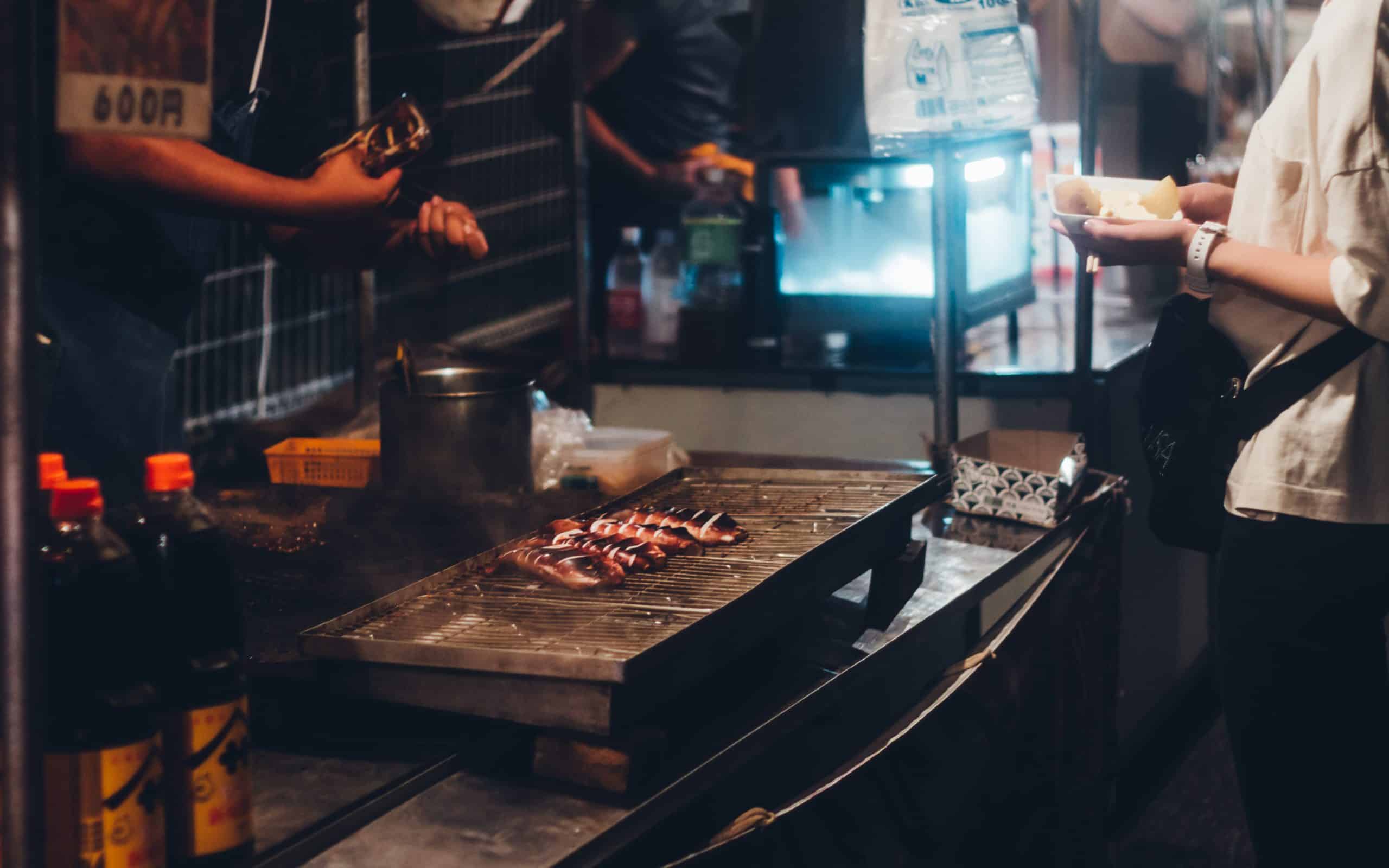 A lit-up food stall at a Japanese summer festival at night, the kind of spot where visitors stand to eat rather than walking away with the food