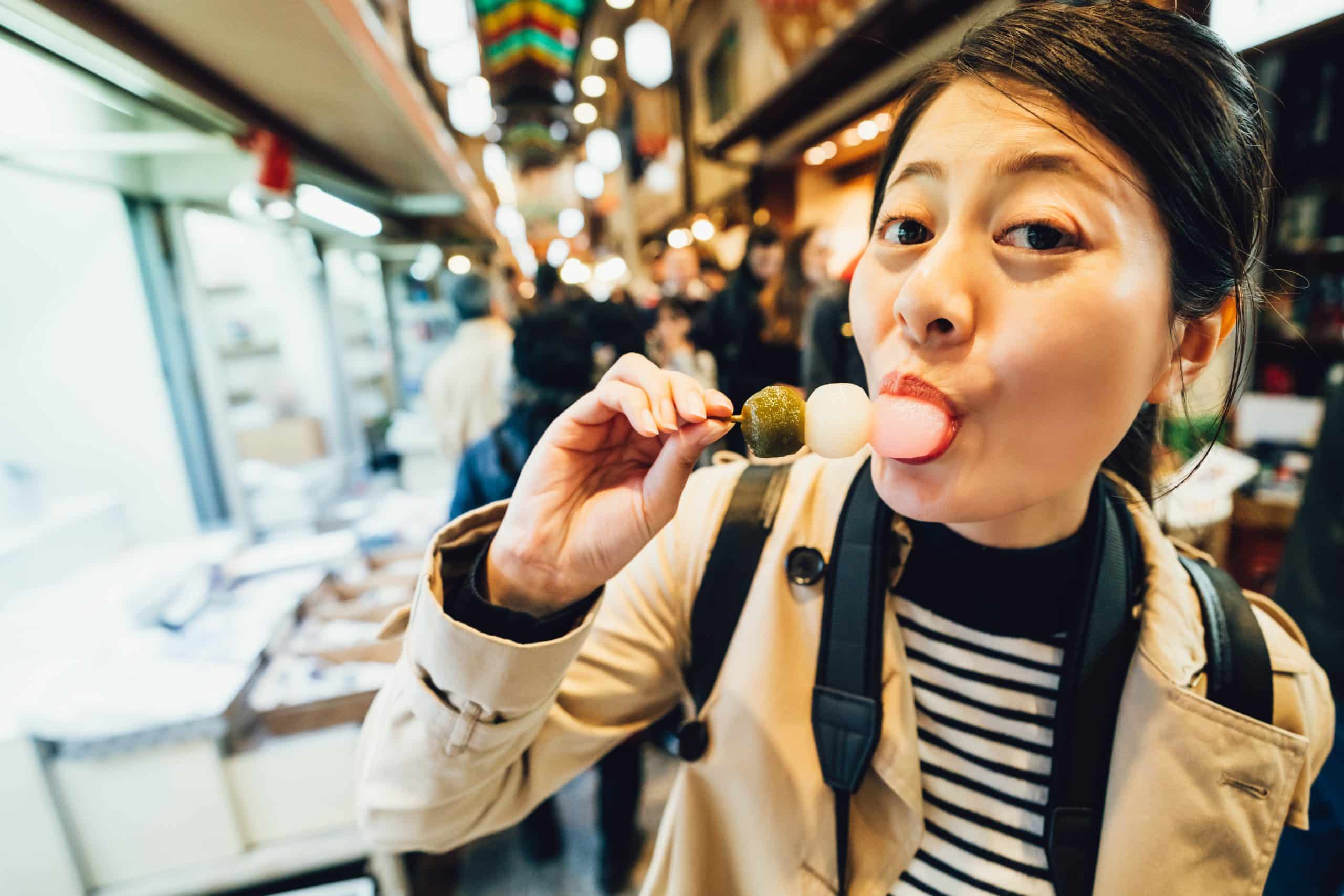 A visitor standing still in front of a vendor stall in Kyoto's Nishiki Market to eat a skewer of dango, rather than eating while walking down the covered arcade